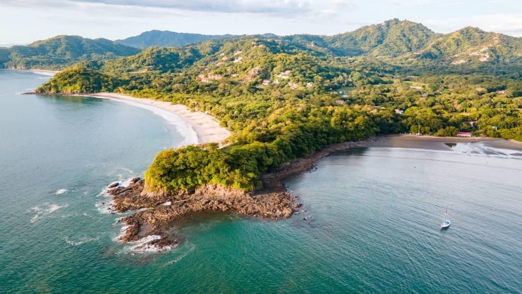An aerial view captures a lush, green peninsula in Costa Rica, edging into a calm blue ocean. This serene setting is home to some of the best beaches in Costa Rica, with sandy shores on both sides and hills rising under a partly cloudy sky.